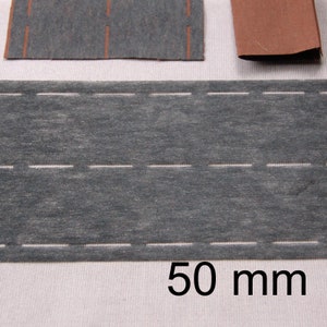 Puede incluir: Una tira de papel gris texturizado con dos líneas blancas discontinuas, que se asemeja a una carretera. La imagen también muestra trozos más pequeños de material similar, uno con líneas verticales naranjas y otro trozo marrón. El texto "50 mm" se muestra en negro.