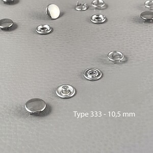 Pode incluir: Vários fechos de pressão de metal prateado, tipo 333, numa superfície cinzenta. Cada fecho, com cerca de 10,5 mm de diâmetro, é composto por uma tampa e um encaixe. A imagem mostra os fechos de diferentes ângulos. Texto: Tipo 333 - 10,5 mm.