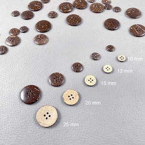 Könnte beinhalten: Eine Auswahl an braunen und beigefarbenen Kokosnussknöpfen in verschiedenen Größen, von 10 mm bis 25 mm. Einige Knöpfe sind dunkelbraun, andere heller. Die Knöpfe haben vier Löcher und liegen auf einer grauen Oberfläche.