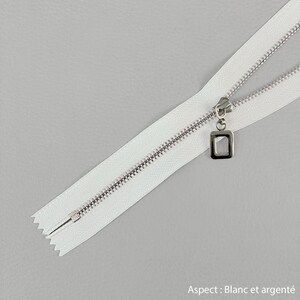 Pode incluir: Fecho branco com dentes e puxador em metal prateado. O puxador tem um design quadrado. O texto "Aspect : Blanc et argenté" é visível no canto inferior direito.