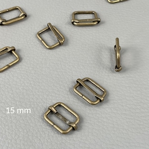 Pode incluir: Deslizadores de metal com cor de latão antigo, com 15 mm, espalhados sobre uma superfície cinza. Os deslizadores são retangulares com uma barra central e uma borda arredondada. A imagem mostra vários deslizadores.