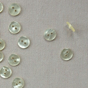 Può includere: Una collezione di bottoni rotondi traslucidi con due fori ciascuno, sparsi su un tessuto grigio chiaro strutturato. I bottoni hanno una lucentezza madreperlacea. Ideale per cucito e progetti artigianali.
