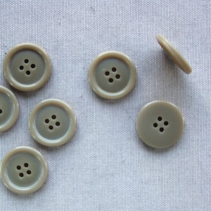 Pode incluir: Vários botões redondos castanhos claros com quatro furos, espalhados sobre um tecido cinza claro. Cada botão tem uma borda externa em relevo. Os botões são de cor neutra, adequados para vários projetos de costura. Diâmetro de cerca de 2,5 cm.