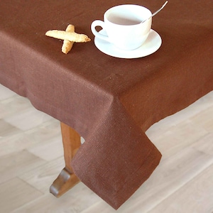 Puede incluir: Un mantel de lino marrón cubre una mesa de madera. Una taza de té blanca con un platillo y una cucharita están sobre el mantel. Dos galletas biscotti están colocadas sobre la mesa cerca de la taza.