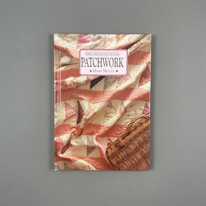 Könnte beinhalten: Ein Buch mit dem Titel "Patchwork" von Mary Moody, Teil der "Serie Artisanat Rural". Das Cover zeigt einen Quilt mit rosa und weißem Stoff und einem Weidenkorb. Das Buch liegt auf einer grauen Oberfläche.