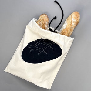 May include: A cream-colored canvas bread bag with a black bread graphic. Two baguettes are visible, protruding from the top. The bag has a drawstring closure with a black cord. The bag is on a gray surface.