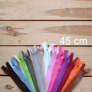 Puede incluir: Una colección de cremalleras de 45 cm de colores variados dispuestas sobre un fondo de madera clara. Las cremalleras son de diversos colores, incluyendo negro, azul, verde, gris, blanco, rosa, morado, naranja y marrón. Se muestra el texto "45 cm".