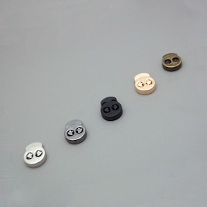 May include: An assortment of cord locks in various metallic finishes, including silver, black, gold, and bronze. Each cord lock features two holes for cord insertion and a spring-loaded mechanism for secure fastening. These small, functional components are used in apparel and accessories.