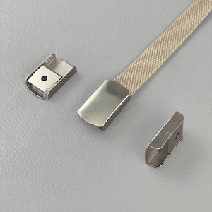 Puede incluir: Un cinturón tejido de color beige con hebilla y terminales de metal plateado. Se muestra sobre un fondo gris claro.