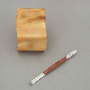 Puede incluir: Un bloque de jabón marrón claro con vetas más oscuras junto a un cuchillo pequeño. El cuchillo tiene un mango de madera y una hoja plateada. El jabón tiene una forma ligeramente irregular.