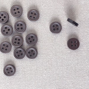 Puede incluir: Un conjunto de botones marrón oscuro de cuatro agujeros esparcidos sobre una tela gris claro texturizada. Los botones son redondos con un borde ligeramente elevado y cuatro agujeros espaciados. Un botón está de lado.