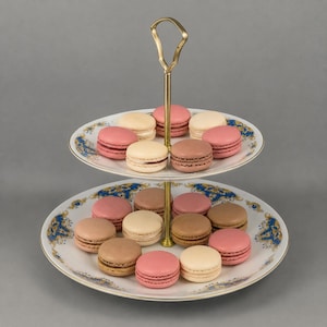 May include: A two-tiered cake stand with a gold handle, displaying a selection of colourful macarons. The macarons, in pink, brown, and cream, are arranged on plates with a blue and gold floral pattern.