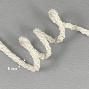 May include: Close-up of a length of off-white, braided rope, approximately 8 mm in diameter. The rope is looped and curved, with a textured appearance. The background is a solid gray surface.