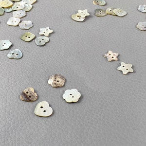 Puede incluir: Una colección de botones decorativos en varias formas, incluyendo cuadrados, corazones, estrellas y flores. Los botones son en tonos de blanco, crema y marrón, con dos o cuatro agujeros para coser. Los botones están dispersos sobre una superficie gris.