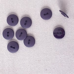 Pode incluir: Uma coleção de oito botões redondos roxos escuros, cada um com dois furos, e um botão com haste. Os botões estão espalhados sobre um tecido bege claro. Os botões são feitos de um material liso e fosco.