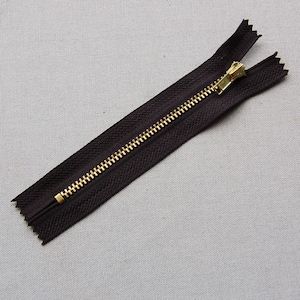 Pode incluir: Um fecho de correr castanho escuro com um puxador e dentes de metal dourado. O fecho de correr está aberto, mostrando os dentes. A fita de tecido é de cor castanho escuro liso.