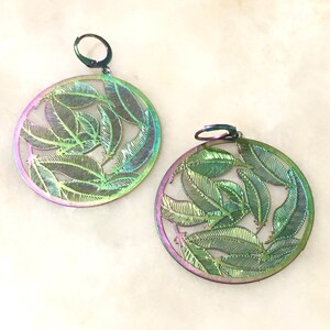 Puede incluir: Un par de pendientes de color verde y morado iridiscente con un diseño circular con hojas recortadas.