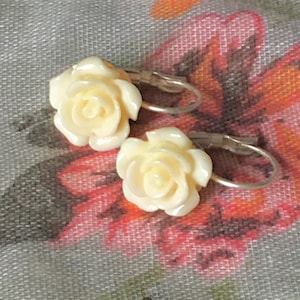 Könnte beinhalten: Cremefarbene Rosenohrringe mit silberfarbenen Bügelverschlüssen. Die Ohrringe weisen detaillierte, dreidimensionale Rosendesigns auf. Der Hintergrund ist ein Blumenmuster in Rosa-, Orange- und Grautönen.