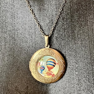 May include: A vintage-style brass locket necklace featuring a round pendant with a colorful hot air balloon design. The pendant has a floral border and hangs from a delicate chain. The locket is closed.