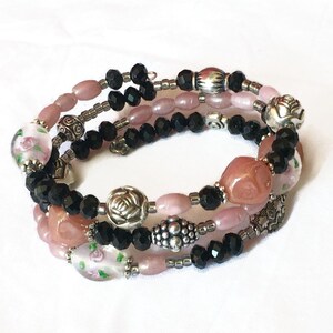 Op de afbeelding: Een multi-strand wikkelarmband met een mix van kralen. De armband bevat zwarte facetkralen, roze ovale kralen en decoratieve zilverkleurige metalen kralen. Sommige kralen hebben een bloemenpatroon.
