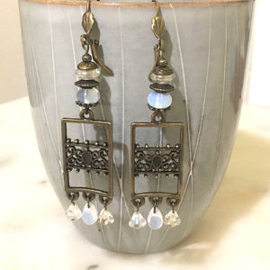 Pode incluir: Um par de brincos de bronze com um design de filigrana e contas de opala branca. Os brincos têm um design pendente com três pequenas contas de cristal na parte inferior.