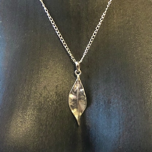 Op de afbeelding: Een zilveren ketting met een bladvormige hanger. De hanger is getextureerd en heeft een licht gebogen vorm.