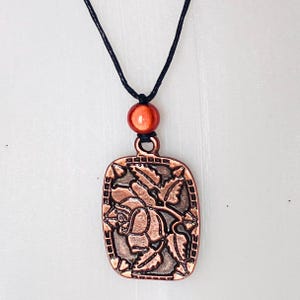 Può includere: Collana con pendente color rame con un pendente rettangolare con un disegno di rosa. Il pendente è sospeso a un cordino nero e include una piccola perla rotonda arancione. La rosa è dettagliata con foglie e steli.