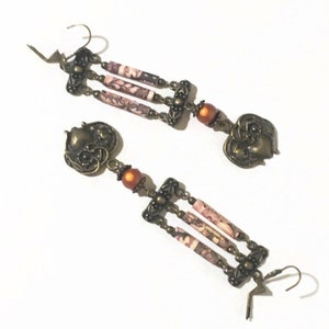 Peut inclure: Une paire de longues boucles d'oreilles pendantes avec un charme en forme de cœur en bas. Les boucles d'oreilles sont en métal brun et ont des perles à motifs roses et blancs.