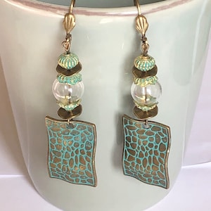 Könnte beinhalten: Ein Paar Hängeohrringe mit türkisfarbener und bronzefarbener Oberfläche. Die Ohrringe haben eine klare Glasperle, einen Stapel aus Bronze- und Türkisperlen und einen quadratischen Anhänger mit einer strukturierten Oberfläche.