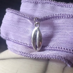 Peut inclure: Un charme en forme de coquille marine argentée pend à un ruban lavande. Le ruban est plié sur lui-même, créant un effet de superposition.