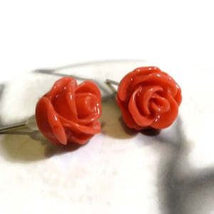 Può includere: Un paio di orecchini a bottone a forma di rosa rossa corallo. Gli orecchini sono realizzati in un materiale liscio e lucido e hanno un design delicato e femminile.