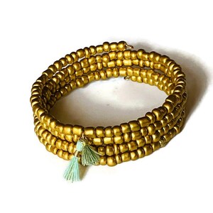 Può includere: Bracciale avvolgente dorato con perline quadrate. Presenta un design a spirale ed è adornato da due piccoli nappe verde chiaro. Il bracciale è progettato per avvolgersi più volte attorno al polso.