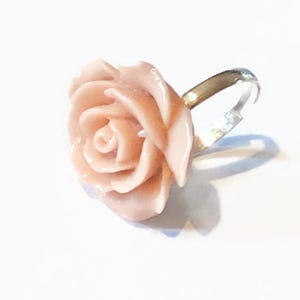 Könnte beinhalten: Ein hellrosa rosenförmiger Ring mit einem verstellbaren Silberband. Die Rose hat detaillierte Blütenblätter und ein glänzendes Finish, vor einem weißen Hintergrund. Das Schmuckstück ist ein Accessoire mit floralem Thema.