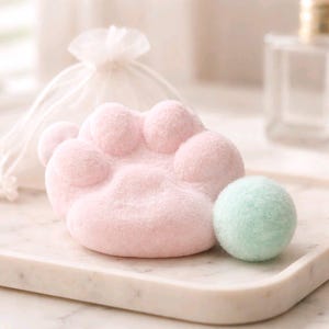 Puede incluir: Una bomba de baño rosa con forma de pata y una pequeña bomba de baño verde claro se encuentran en una bandeja de mármol. Una bolsa transparente con cordón blanco está en el fondo. Las bombas de baño tienen un aspecto suave y texturizado.