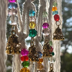 May include: A decorative macrame wall hanging featuring white rope strands. The strands are adorned with colourful faceted beads in pink, blue, green, yellow, and orange. Gold-coloured bell charms hang at intervals, adding a touch of elegance.