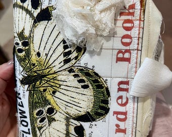 Könnte beinhalten: Ein handgefertigtes Stoffjournal mit Schmetterlingsdesign. Das Cover zeigt eine große Schmetterlingsillustration in Schwarz, Gelb und Grün, mit den Worten "Flower Garden Book" in Rot. Eine weiße Spitzenblume mit Perlenmitte ziert die Oberseite.