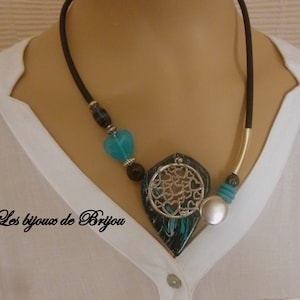 Op de afbeelding: Een zwarte en turquoise ketting met een grote hanger met een zilveren hartontwerp. De hanger is gemaakt van een gemarmerd materiaal met een zwart en turquoise kleurenschema. De ketting is gemaakt van zwart koord en heeft een gouden accent.