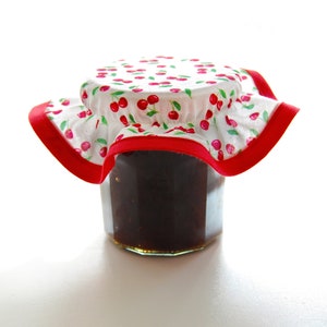 Può includere: Un barattolo di vetro pieno di marmellata marrone scuro, sormontato da un coperchio in tessuto bianco con un bordo rosso e una stampa di ciliegie.
