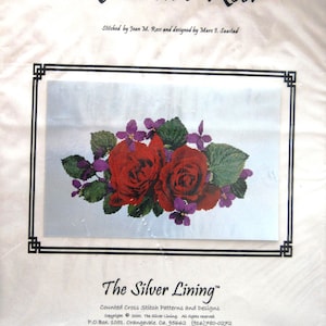 May include: A cross-stitch pattern titled "Roses are Red" featuring two red roses surrounded by green leaves and purple flowers. The design is framed with a black border and includes the text "The Silver Lining" and designer information.