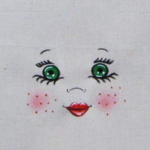 Puede incluir: Una cara de muñeca pintada a mano con ojos verdes, boca roja y mejillas rosadas sobre un fondo de tela blanca.