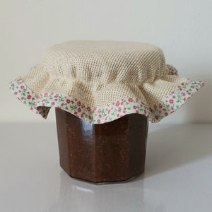 Può includere: Un barattolo di vetro pieno di una sostanza marrone scuro, sormontato da un coperchio in tessuto beige con un bordo floreale rosa e verde.