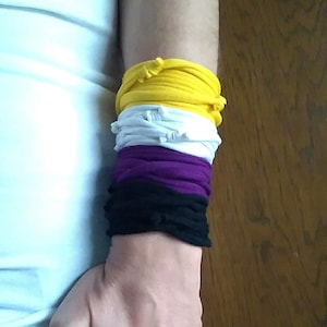 Könnte beinhalten: Ein Arm mit einem Stapel Stoffarmbänder in den Farben Gelb, Weiß, Lila und Schwarz. Die Person trägt eine blaue Jeans.