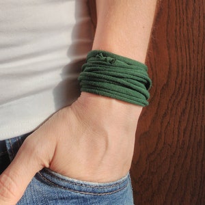 Può includere: Una persona indossa una canottiera bianca e jeans blu con un braccialetto in tessuto verde scuro al polso.