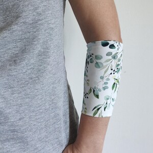 Puede incluir: Una persona lleva una camiseta gris y pantalones vaqueros azules con una banda blanca para el brazo con un patrón de hojas de eucalipto verdes. El texto "Eucalyptus" es visible en la parte inferior de la imagen.