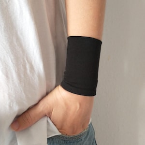 Peut inclure: Un bracelet noir en tissu doux et extensible. Le bracelet est porté au poignet gauche d'une personne portant une chemise blanche et un jean bleu. Le mot "Noir" est écrit en blanc sous le poignet.