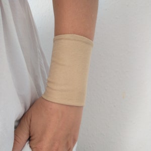 Könnte beinhalten: Ein beiges Armband aus geripptem Strickstoff. Das Armband wird am Arm getragen und bedeckt den Handgelenkbereich. Die Person trägt ein weißes Hemd und eine blaue Jeans.