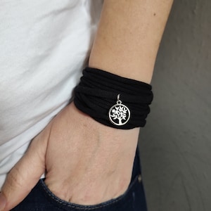 May include: A black fabric bracelet with a silver tree of life charm. The bracelet is wrapped around a person's wrist.
