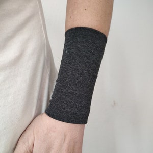 Op de afbeelding: Een armwarmer in antracietgrijs gemaakt van een zachte, rekbare stof. De armwarmer wordt gedragen op de linkerarm van een persoon.