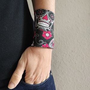 Pu&ograve; includere: Un braccialetto nero con un designo di teschio di zucchero colorato. Il teschio ha dettagli rosa e bianchi ed &egrave; circondato da fiori rosa e viti nere.