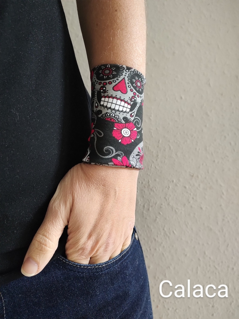 Pu&ograve; includere: Un braccialetto nero con un designo di teschio di zucchero colorato. Il teschio ha dettagli rosa e bianchi ed &egrave; circondato da fiori rosa e viti nere.